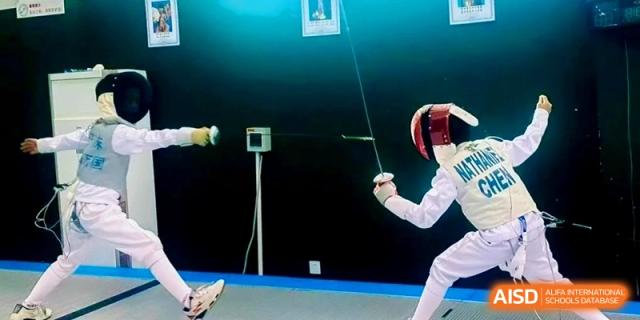 Two students engage in a competitive fencing match on a blue piste, wearing full protective gear and wielding foils at Shanghai Qingpu World Foreign Language High School.