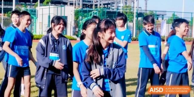 A group of smiling students in blue uniforms and grey jackets shares a cheerful moment together on the grassy field at Shanghai Qingpu World Foreign Language High School.