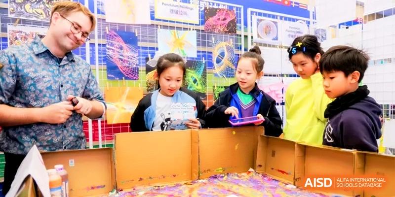 Two girls stand behind a cardboard display showcasing colorful geometric art projects at Shanghai United International School, Hongqiao Campus.