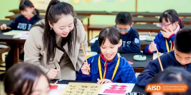 A teacher guides a student through a calligraphy or art project at a wooden desk. | Shanghai United International School, Hongqiao Campus