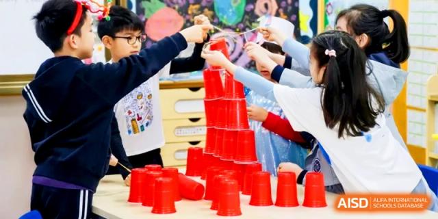 A group of students collaborates to build a tall tower using many red plastic cups. | Shanghai United International School, Hongqiao Campus