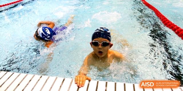 A young student wearing goggles and a blue swim cap enjoys a swimming lesson in the clear blue water of the indoor pool at Shanghai United International School, Hongqiao Campus