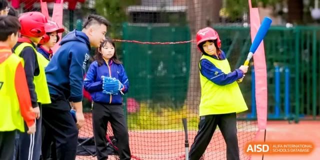 Students wearing safety helmets and athletic gear participate in a baseball or softball practice session on the field at Shanghai United International School, Hongqiao Campus