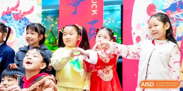 Two young students in festive attire make heart shapes with their hands during a celebration at Shanghai United International School, Hongqiao Campus