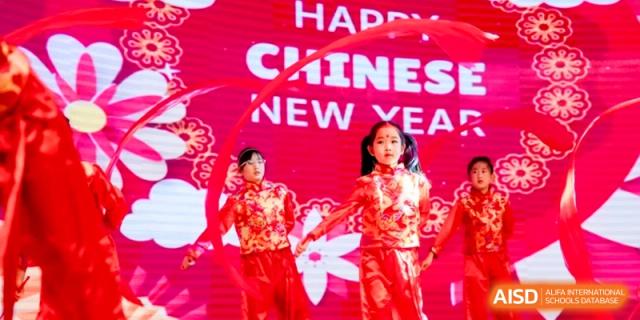 Young girls in traditional red dresses perform a ribbon dance in front of a Happy Chinese New Year sign at Shanghai United International School, Hongqiao Campus