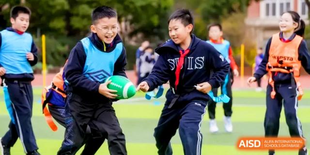 Two students in athletic wear laugh while playing a game of football on a green outdoor field at Shanghai United International School, Hongqiao Campus