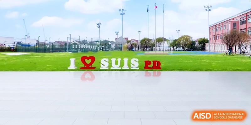 Large outdoor signage reading I Heart SUIS PD stands on a green field under a clear blue sky with flags at Shanghai United International School, Pudong.