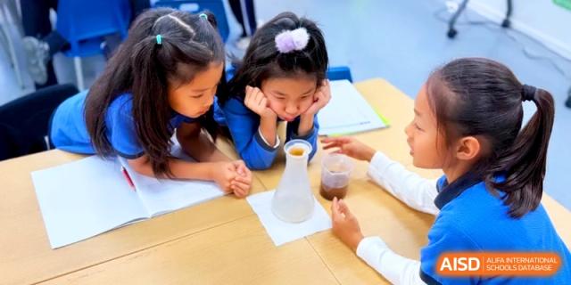 Young students look closely at a glass flask during a hands-on science lesson at a wooden desk at Shanghai United International School, Pudong