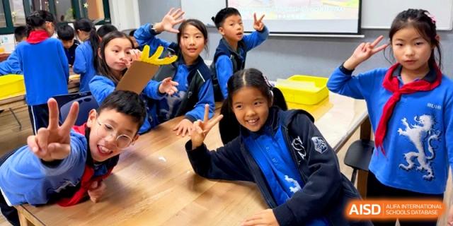 Happy students pose and make peace signs while working on projects at a large wooden desk at Shanghai United International School, Pudong