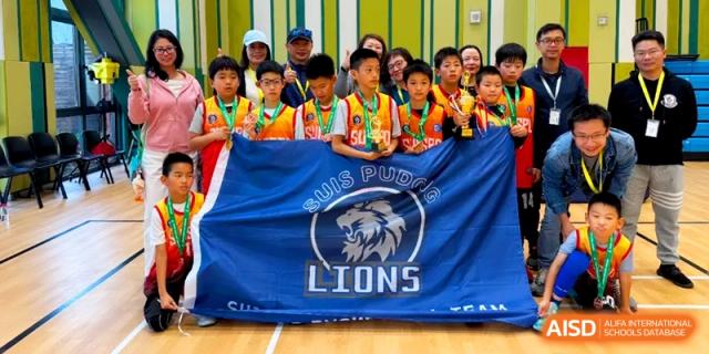 A youth basketball team poses proudly with a large blue SUIS Pudong Lions flag and trophies at Shanghai United International School, Pudong