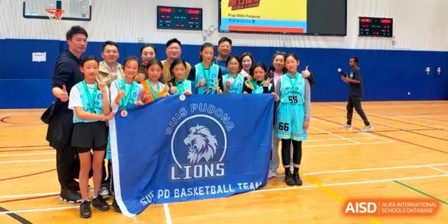 A group of female basketball players poses proudly with a large blue SUIS Pudong Lions flag on an indoor court at Shanghai United International School, Pudong