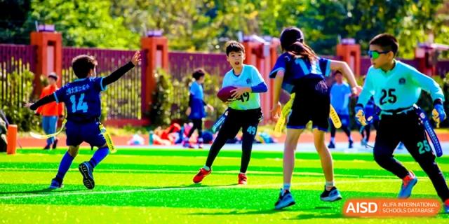 A student athlete wearing a blue jersey carries a football during a flag football game on a green turf field at Shanghai United International School, Pudong