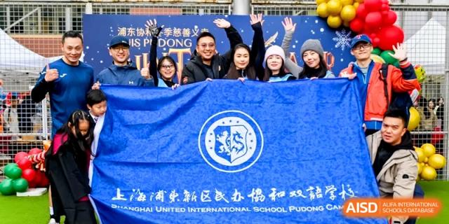 Staff members smile while holding a large blue school banner featuring a white crest during a community celebration at Shanghai United International School, Pudong