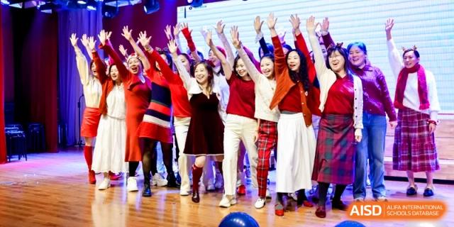 Teachers in festive red and white outfits perform together on a brightly lit stage during a holiday event at Shanghai United International School, Pudong