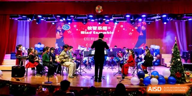 A conductor leads a student wind ensemble performing on a stage with a vibrant pink and purple background at Shanghai United International School, Pudong