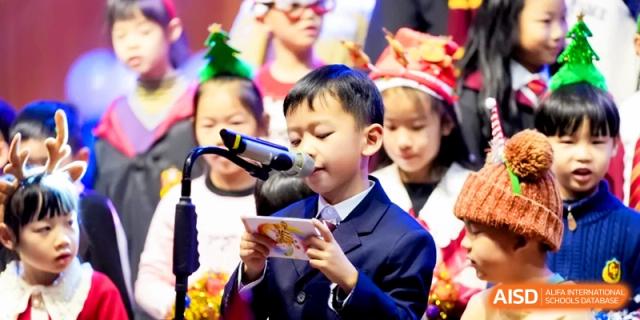 A young boy in a suit speaks into a microphone while holding a script during a formal school assembly or event at Shanghai United International School, Pudong