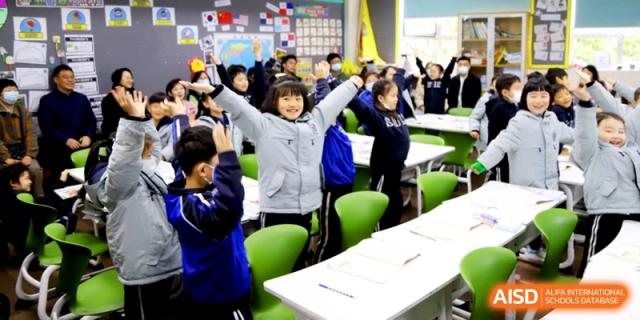 A primary student with arms raised in a cheerful classroom setting during a group activity at Shanghai United International School, Shangyin Campus.