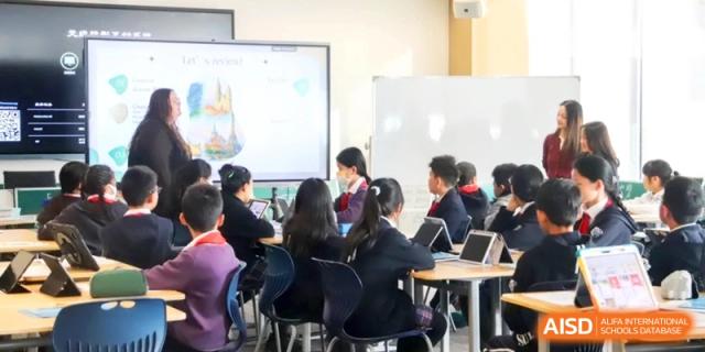 Students at Shanghai United International School, Qingpu Campus sit at desks with tablets while looking at a presentation on a white digital board in a bright, modern classroom setting