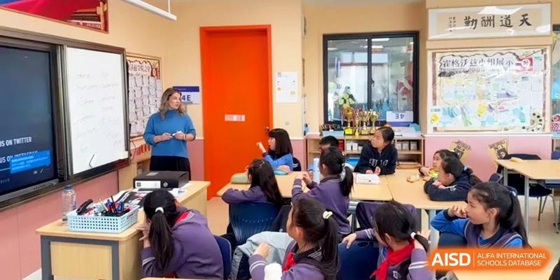 Young students in purple uniforms participate in a classroom discussion led by a teacher standing near a bright orange door at Shanghai United International School, Qingpu Campus