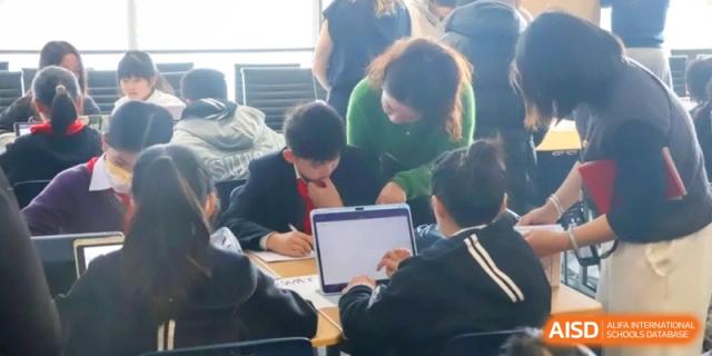 A teacher in a green sweater assists a student working on a laptop during a collaborative lesson at Shanghai United International School, Qingpu Campus with other students nearby