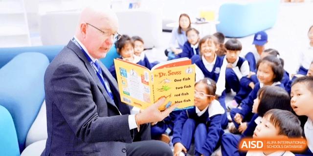 A male teacher reading a Dr. Seuss book to a group of smiling primary students sitting on the floor in a bright classroom at HT Nanjing Impact Academy.