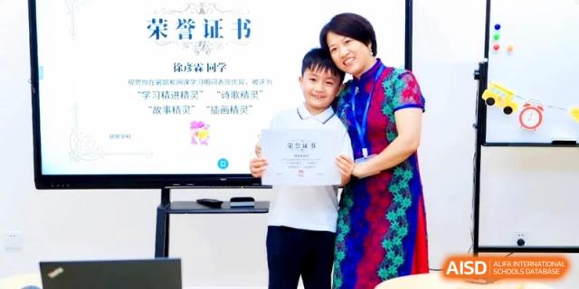 A smiling teacher standing next to a young student who is proudly holding up an award certificate at HT Nanjing Impact Academy.