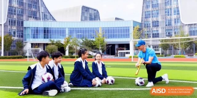 Students sitting on a green turf football field with soccer balls in front of a modern school building at HT Nanjing Impact Academy.