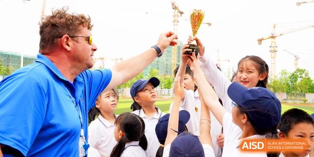 A teacher handing a trophy to a group of excited students celebrating a sports victory outdoors at HT Nanjing Impact Academy.