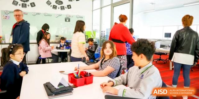 Students in a bright classroom work together at a white table with art supplies while a teacher in red assists nearby at the International School of Tianjin.