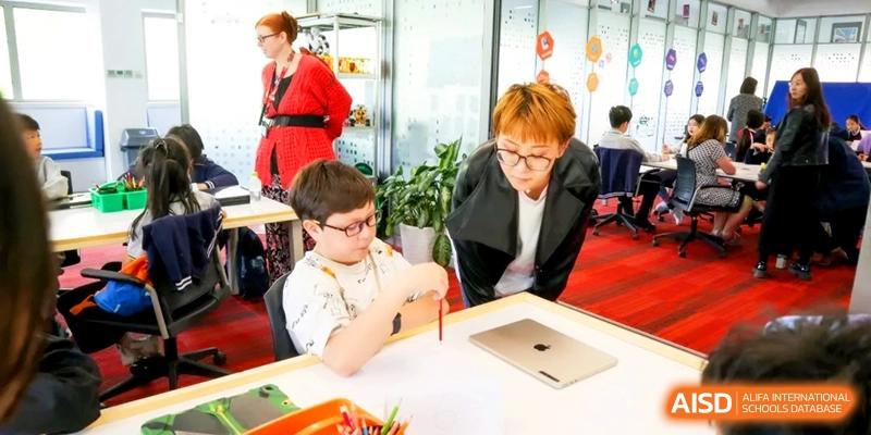 A young student works on a drawing at a desk while a teacher provides individual support at the International School of Tianjin.