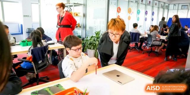 A young student works on a drawing at a desk while a teacher provides individual support at the International School of Tianjin.