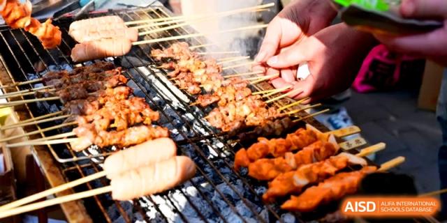 Hands turn rows of seasoned meat skewers and sausages grilling on a large outdoor barbecue at Kunshan Senior High School.