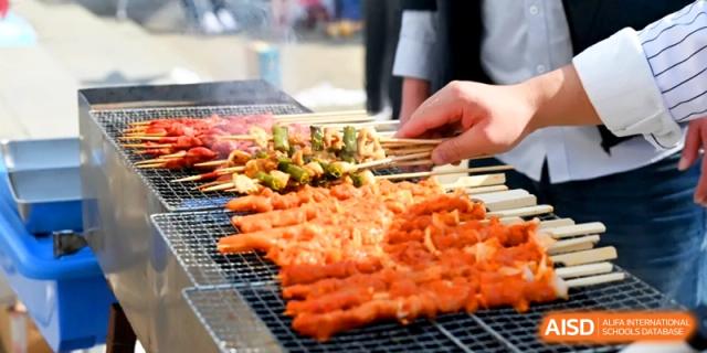 A close-up of various meat and vegetable skewers being grilled over an open flame at a food festival at Kunshan Senior High School.