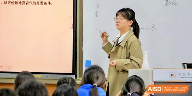 A teacher in a tan coat points to a whiteboard while explaining a complex concept to her class at Ningbo Beilun High School.