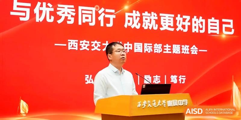 A male speaker in a white shirt stands at a podium giving a presentation in front of a bright red backdrop at The High School Affiliated to Xi_an Jiaotong University.