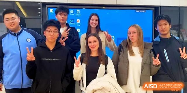 A group of diverse international students smiles for a photo in front of a blue digital screen in a classroom at The High School Affiliated to Xi_an Jiaotong University.