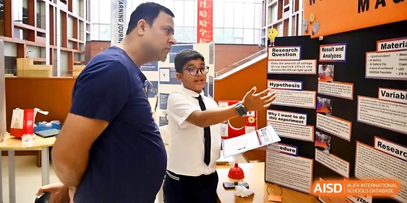 A young student in a tie gestures toward his research poster while explaining a project to an adult at Wahaha International School.