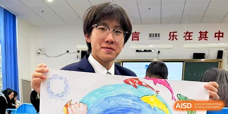 A smiling male student in a suit and glasses holds up a large, colorful painting of a blue globe and fish at WeiFang No.1 Middle School.
