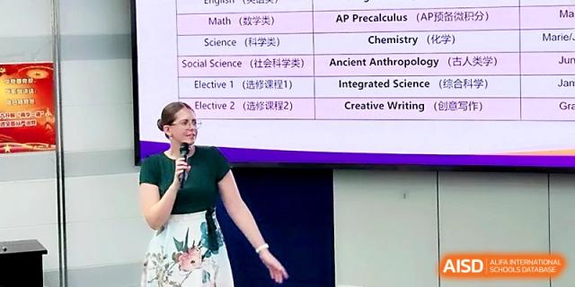 A female teacher gestures toward a presentation slide while speaking into a microphone during an academic seminar at Wuhan No.2 High School.