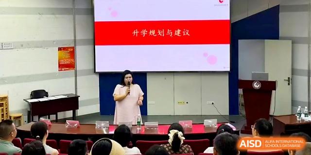 A presenter stands on stage in front of a large screen, delivering an informative lecture to a seated audience at Wuhan No.2 High School.
