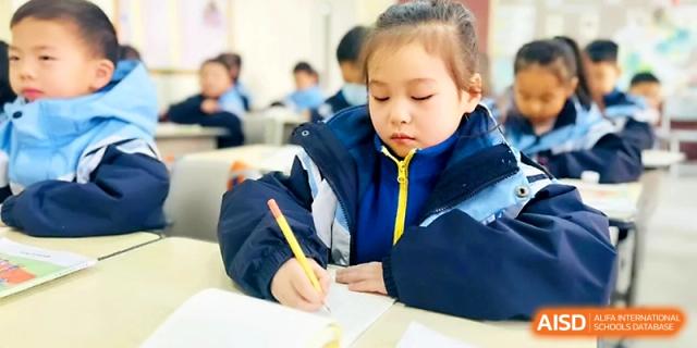 A young girl in a blue winter jacket focuses intently on her writing during a class at Xi_an Bodi School.