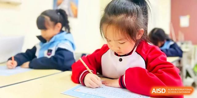 A student in a red and white jacket practices calligraphy or writing at a wooden desk at Xi_an Bodi School.