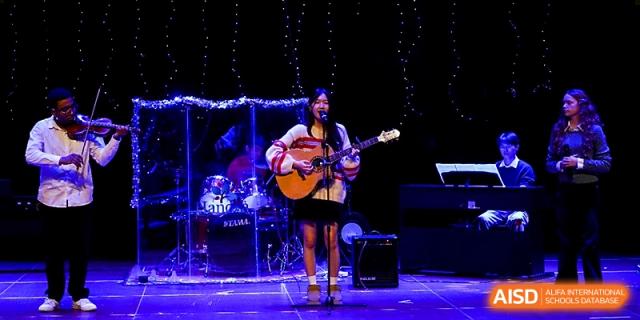 A solo student performer sings while playing an acoustic guitar on a stage with blue lighting at Xi_an Hanova International School.