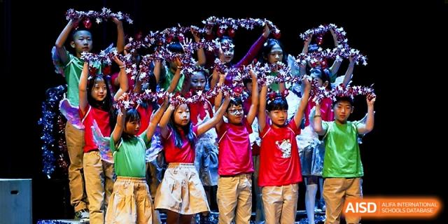 Young students in vibrant red and green shirts hold up pom-poms during a synchronized school performance at Xi_an Hanova International School.