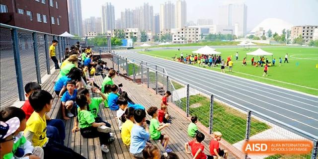 Students in colorful team shirts sit on bleachers overlooking the school running track and field at Xi_an Hanova International School.