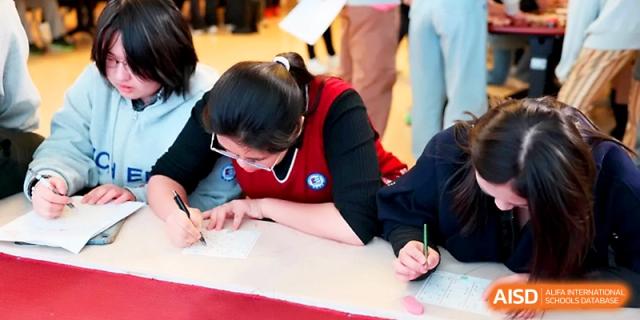 A student in a red vest focuses on writing carefully on a sheet of paper during a classroom activity at Beijing Xicheng Academy.