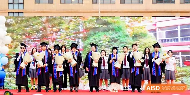 A group of graduates in academic gowns poses together with flower bouquets on campus at Beijing Xicheng Academy.
