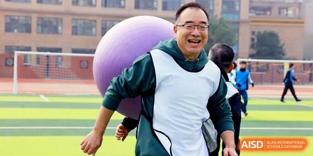 A smiling man participates in a team-building exercise with a large purple fitness ball on the sports field at Beijing Xicheng Academy.