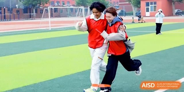 Students in red vests laugh while competing in a partnered race on the green artificial turf field at Beijing Xicheng Academy.