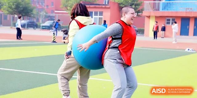 Students wearing red vests work together to move a large blue exercise ball during a team challenge at Beijing Xicheng Academy.